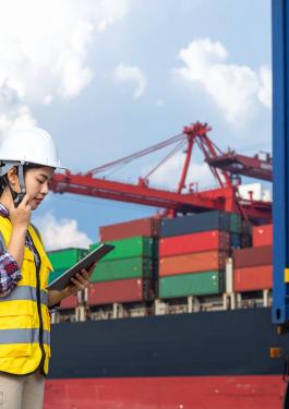 Mujer con chaleco y casco mirando un Tablet frente a barco carguero.